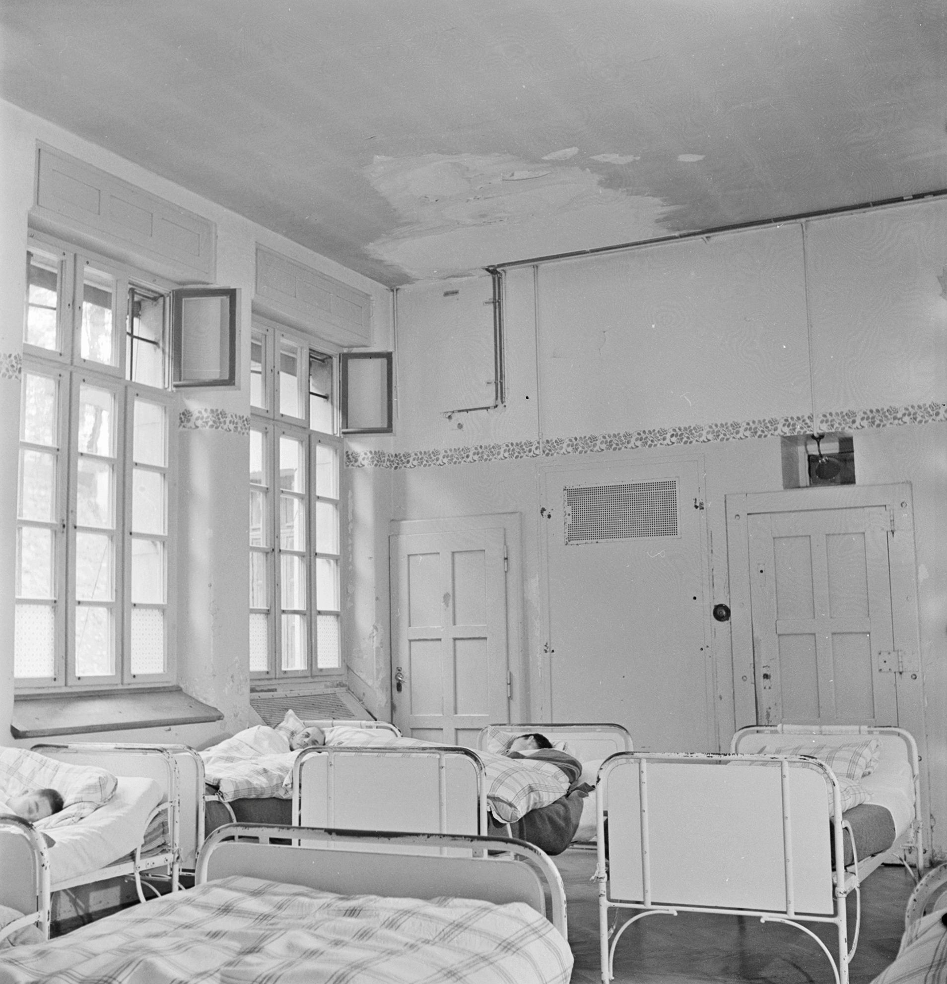 Fotografia in bianco e nero di una stanza in una clinica psichiatrica intorno al 1948. Si vedono diversi letti metallici, tre dei quali occupati da persone che hanno la testa girata dall'altra parte.