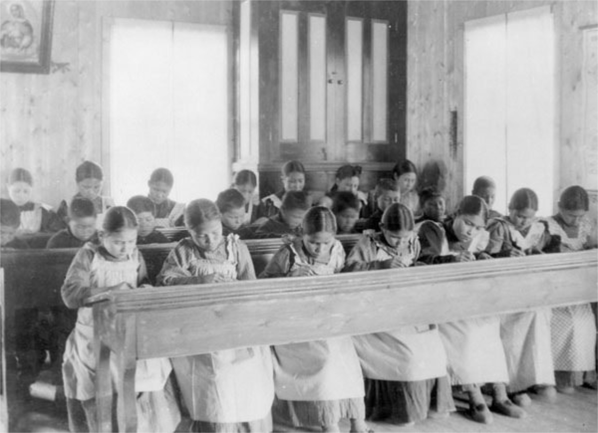 Fotografia in bianco e nero di un'aula scolastica con banchi di legno, dove alcune ragazze sono sedute e scrivono. Si tratta di una cosiddetta “Residential School” (istituto di educazione) per bambini indigeni in Canada.