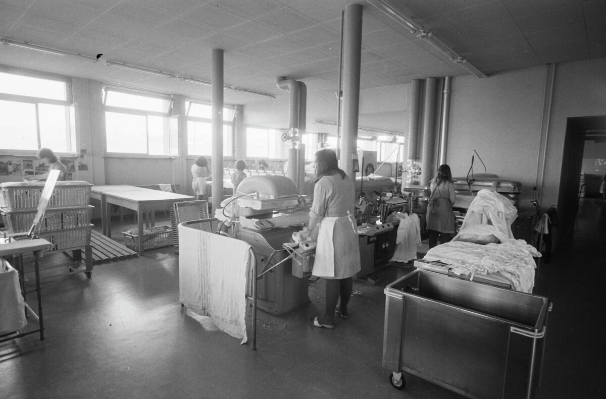 Fotografia in bianco e nero della lavanderia nel carcere femminile di Hindelbank. Si vedono quattro donne di spalle mentre lavorano alle stiratrici. 1967.