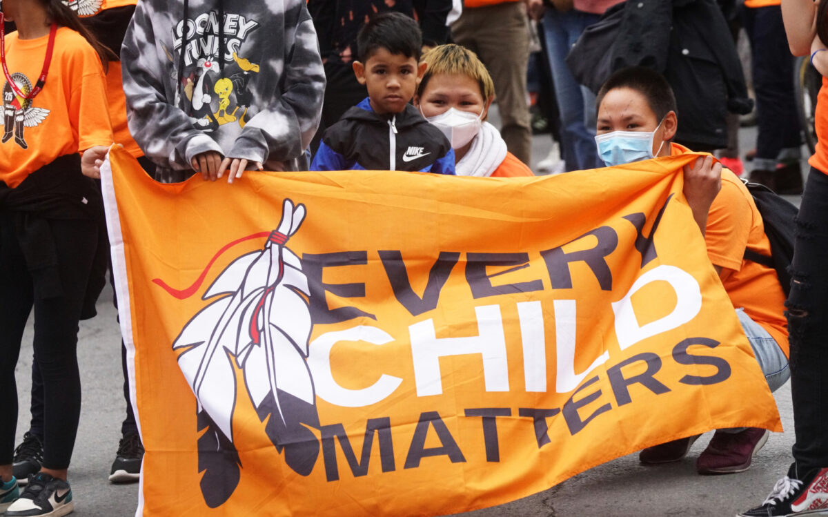 Fotografia a colori di diversi bambini e due adulti dietro uno striscione con la scritta “Every Child Matters”. “Every Child Matters” è un movimento canadese che si concentra sui diritti, il benessere e la dignità di tutti i bambini, in particolare quelli indigeni.