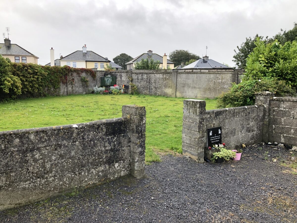 Fotografia a colori di un cortile recintato con muri in pietra. Vista dall'ingresso. All'ingresso e sul lato opposto si vedono fiori e targhe commemorative. Qui si commemorano i bambini morti nella casa madre e figlio Bon Secours a Tuam, Galway.